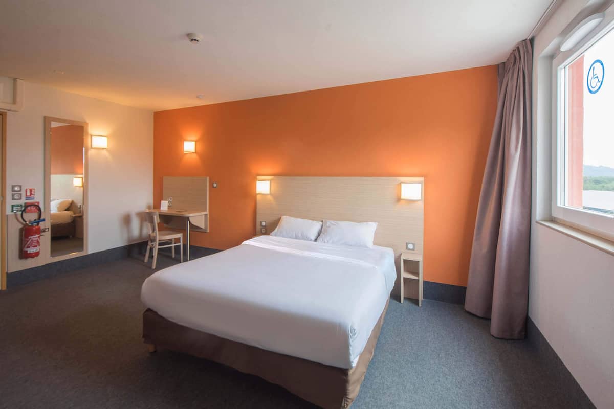 A hotel room with a large bed, a desk, a chair, and a window with curtains.