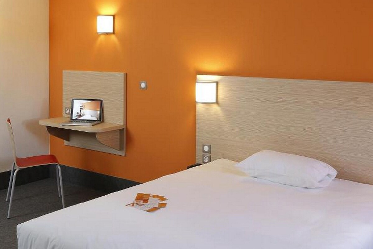 A hotel room with a bed, a desk with a laptop, two wall-mounted lights, and a chair.