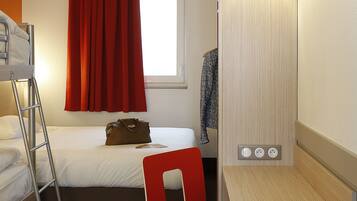 Habitación de hotel con literas, un escritorio con silla, una lámpara y una ventana con cortinas rojas.