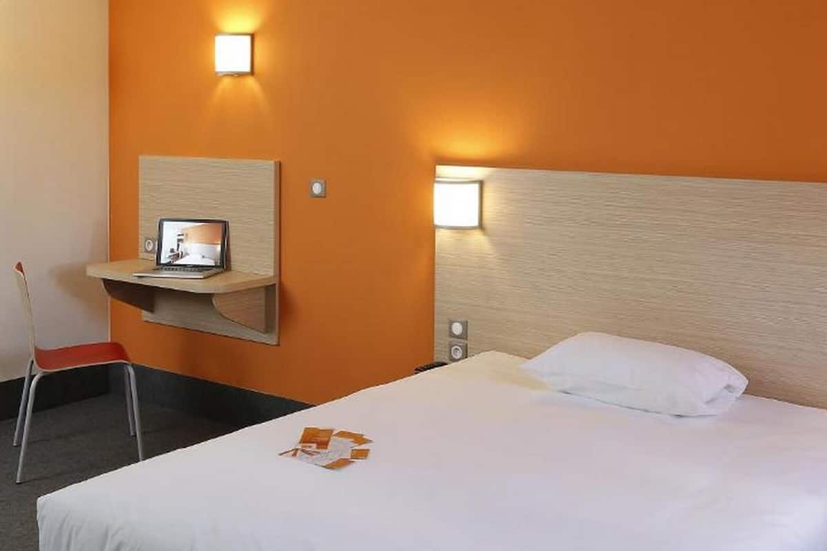 A hotel room with a bed, a desk with a laptop, two wall-mounted lights, and a chair.