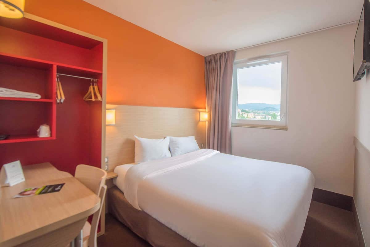 A hotel room with a large bed, a desk with a chair, a red storage unit, and a window with a view.