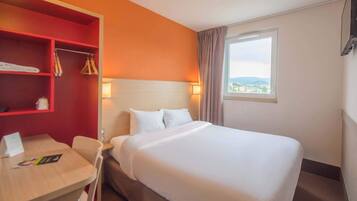 A hotel room with a large bed, a desk with a chair, a red storage unit, and a window with a view.