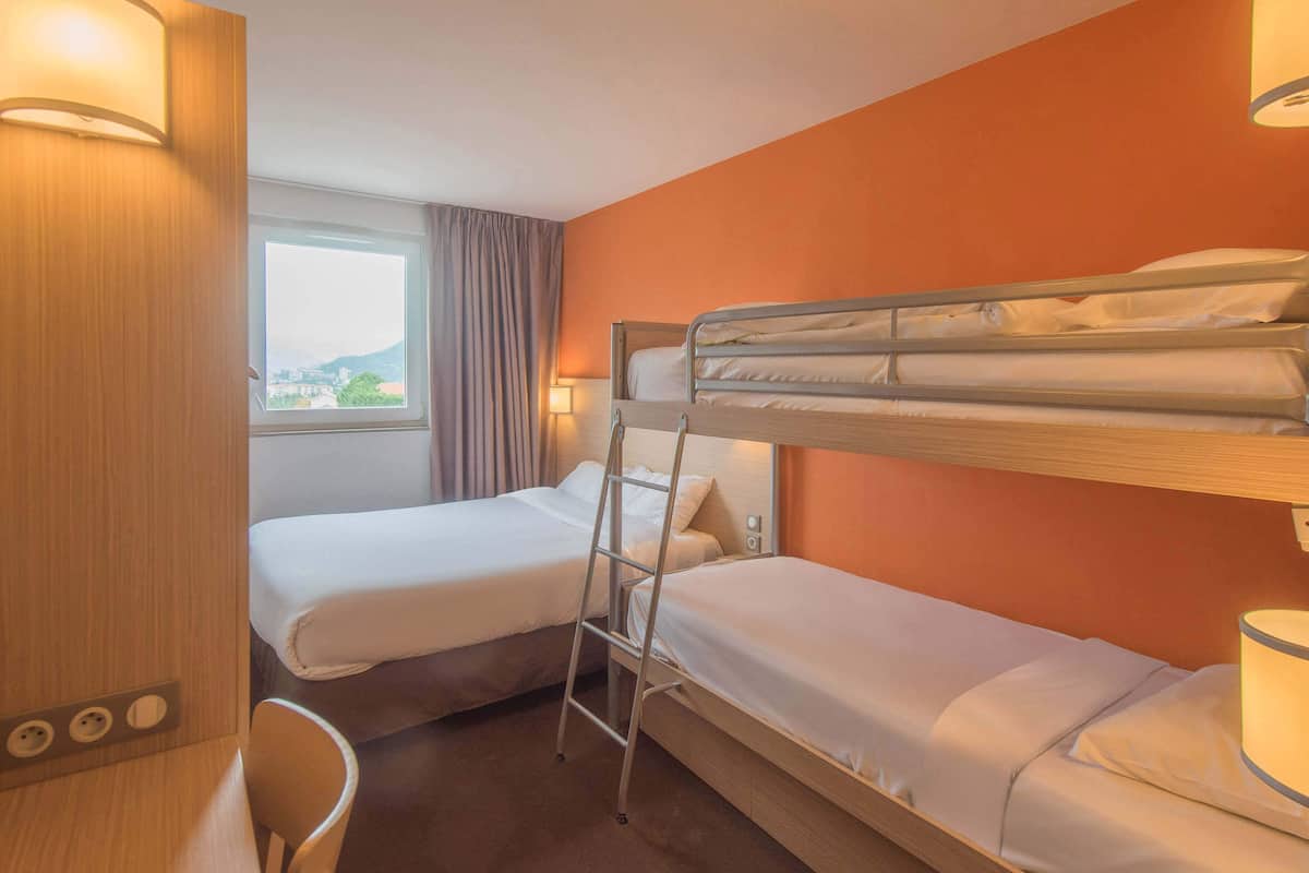 A hotel room with two single beds, a bunk bed, a desk with a chair, a window with curtains, and a lamp.