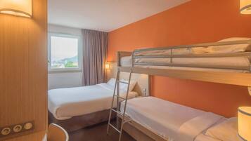 Ein Hotelzimmer mit zwei Einzelbetten, einem Schlafsofa, einem Schreibtisch mit Stuhl, einem Fenster mit Vorhängen und einer Lampe.