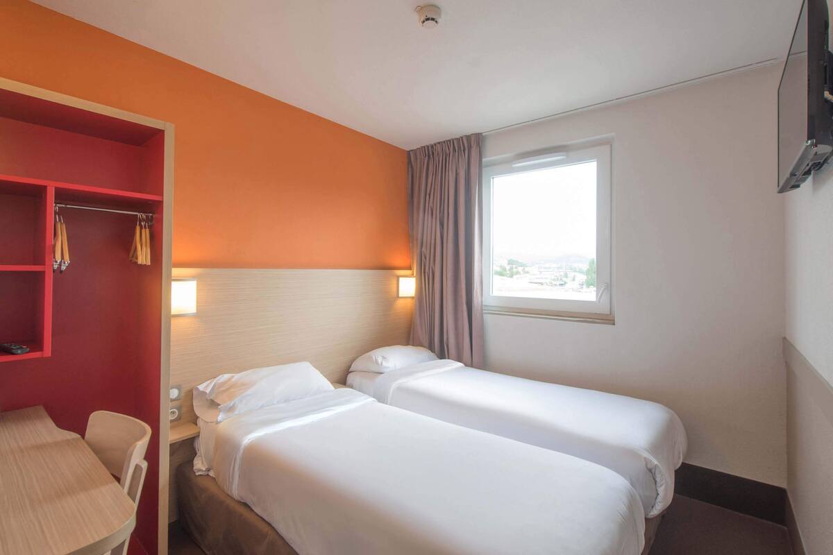 A hotel room with two single beds, a desk, a chair, a red wardrobe, and a window with curtains.