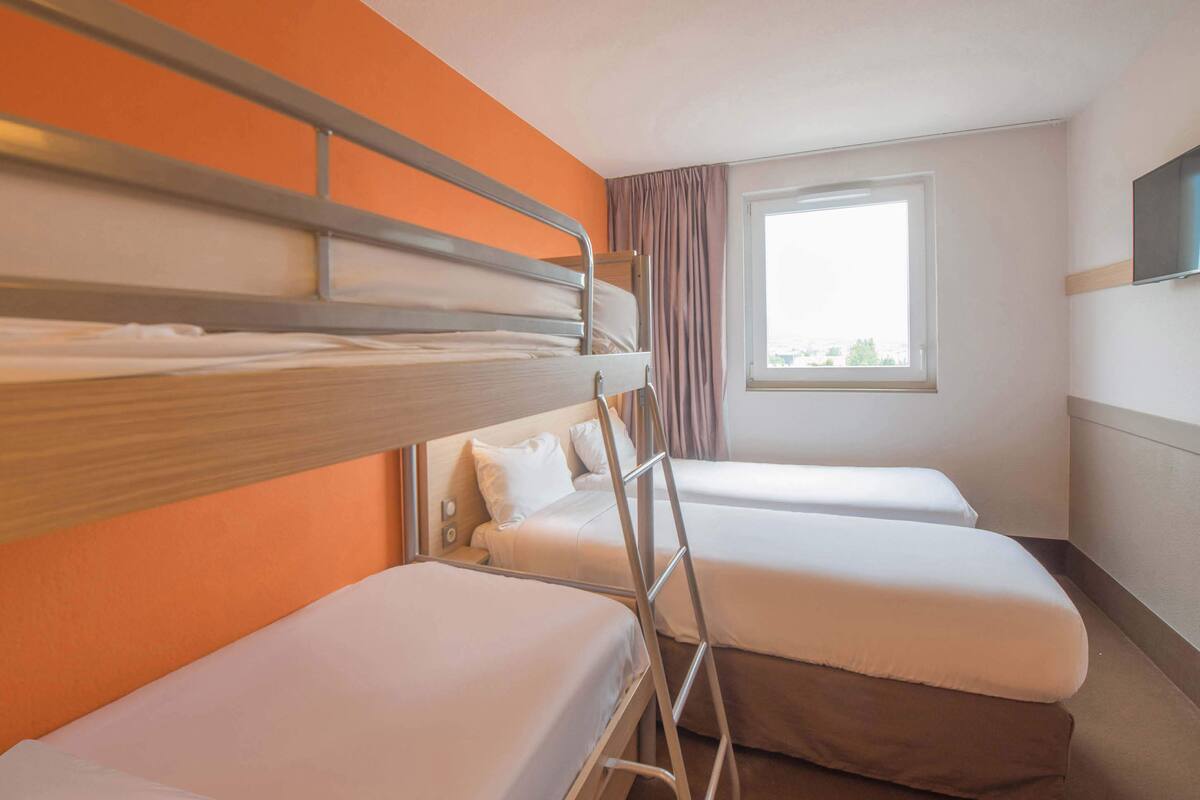 A hotel room with two single beds, a bunk bed, a window with curtains, and a flat-screen TV mounted on the wall.