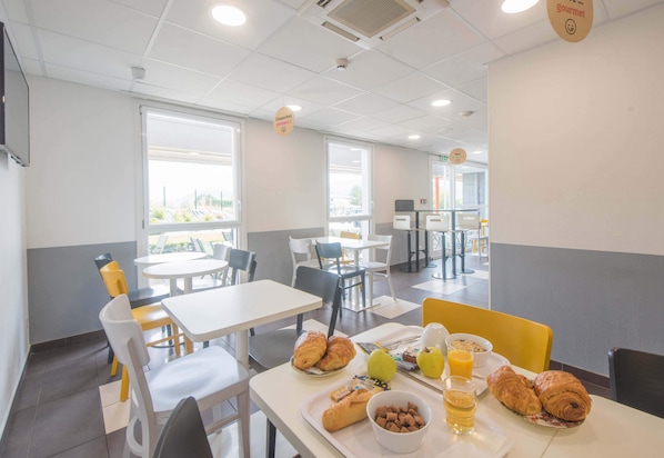 Un espace repas avec des tables et des chaises, des produits pour le petit-déjeuner comme des croissants, des pommes et du jus d’orange, et une vue sur l’extérieur grâce à de grandes fenêtres.
