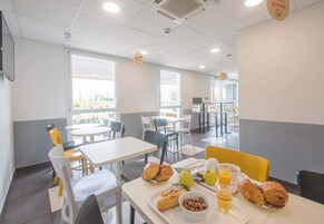 Un espace repas avec des tables et des chaises, des produits pour le petit-déjeuner comme des croissants, des pommes et du jus d’orange, et une vue sur l’extérieur grâce à de grandes fenêtres.