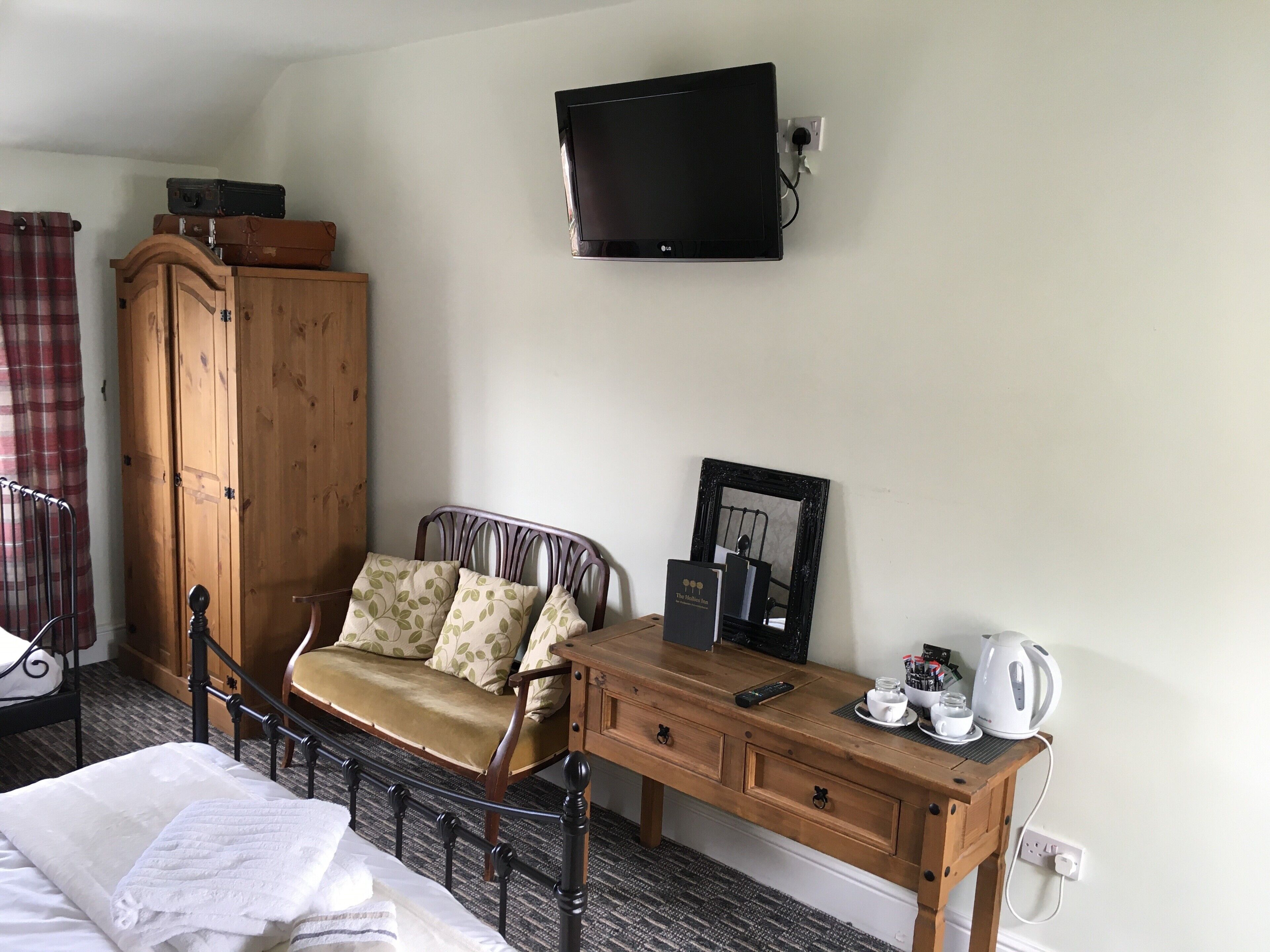 A hotel room with a bed, a wooden desk with a kettle, a chair, a wardrobe, and a flat-screen TV mounted on the wall.
