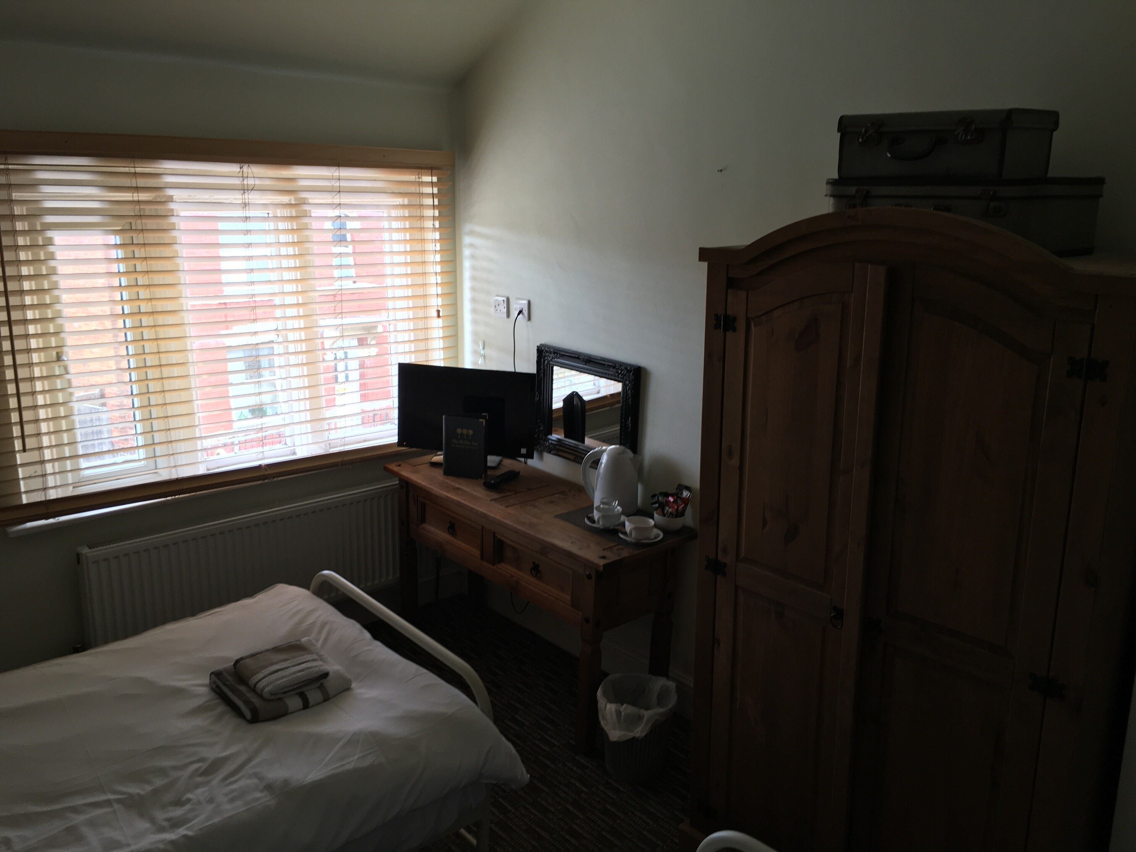 A hotel room with a bed, a desk with a computer, a window with blinds, a radiator, a wooden wardrobe, and a small trash bin.
