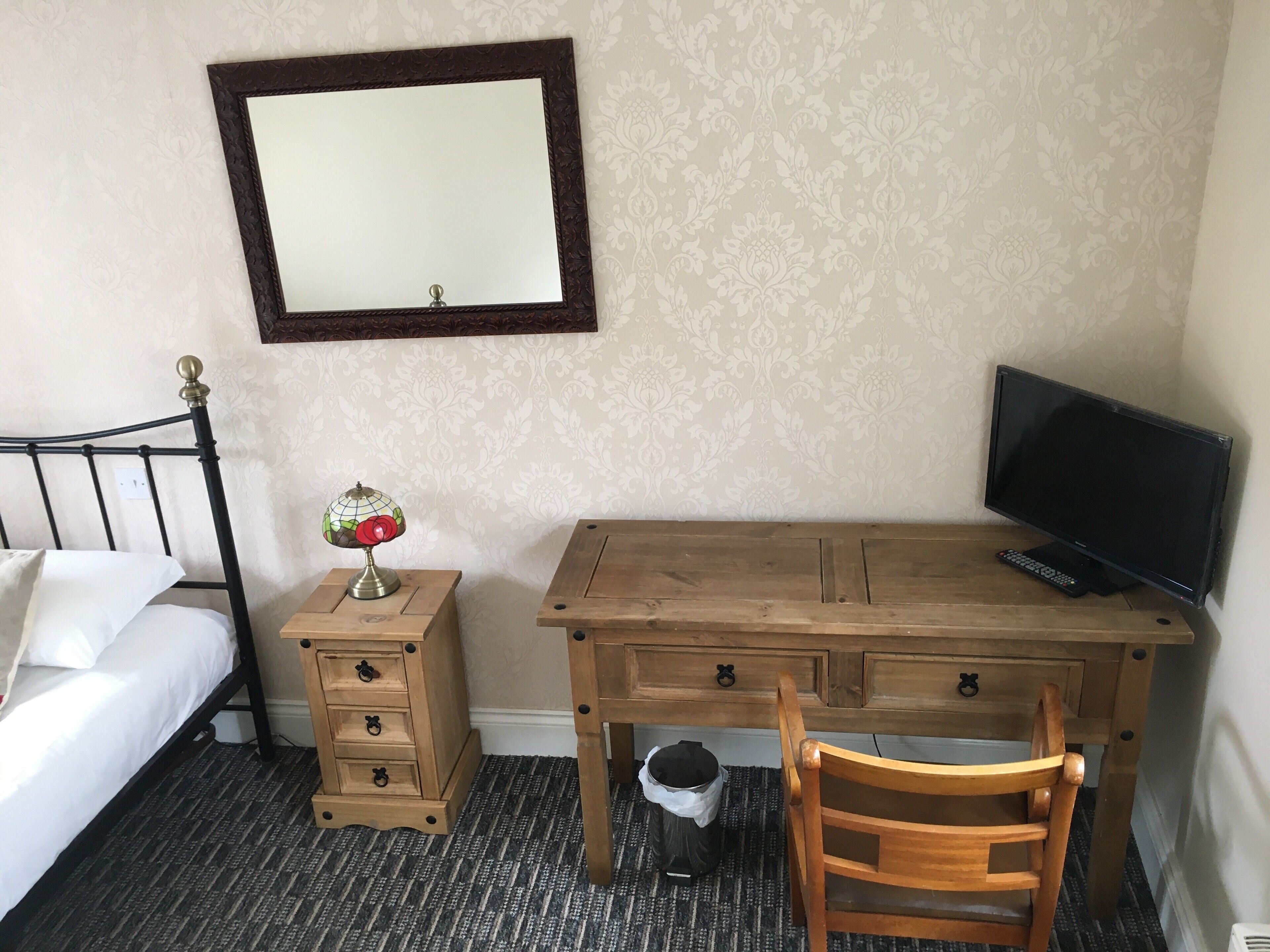 Habitación de hotel con cama, escritorio de madera con televisor, una silla, lámpara y cesto de basura.