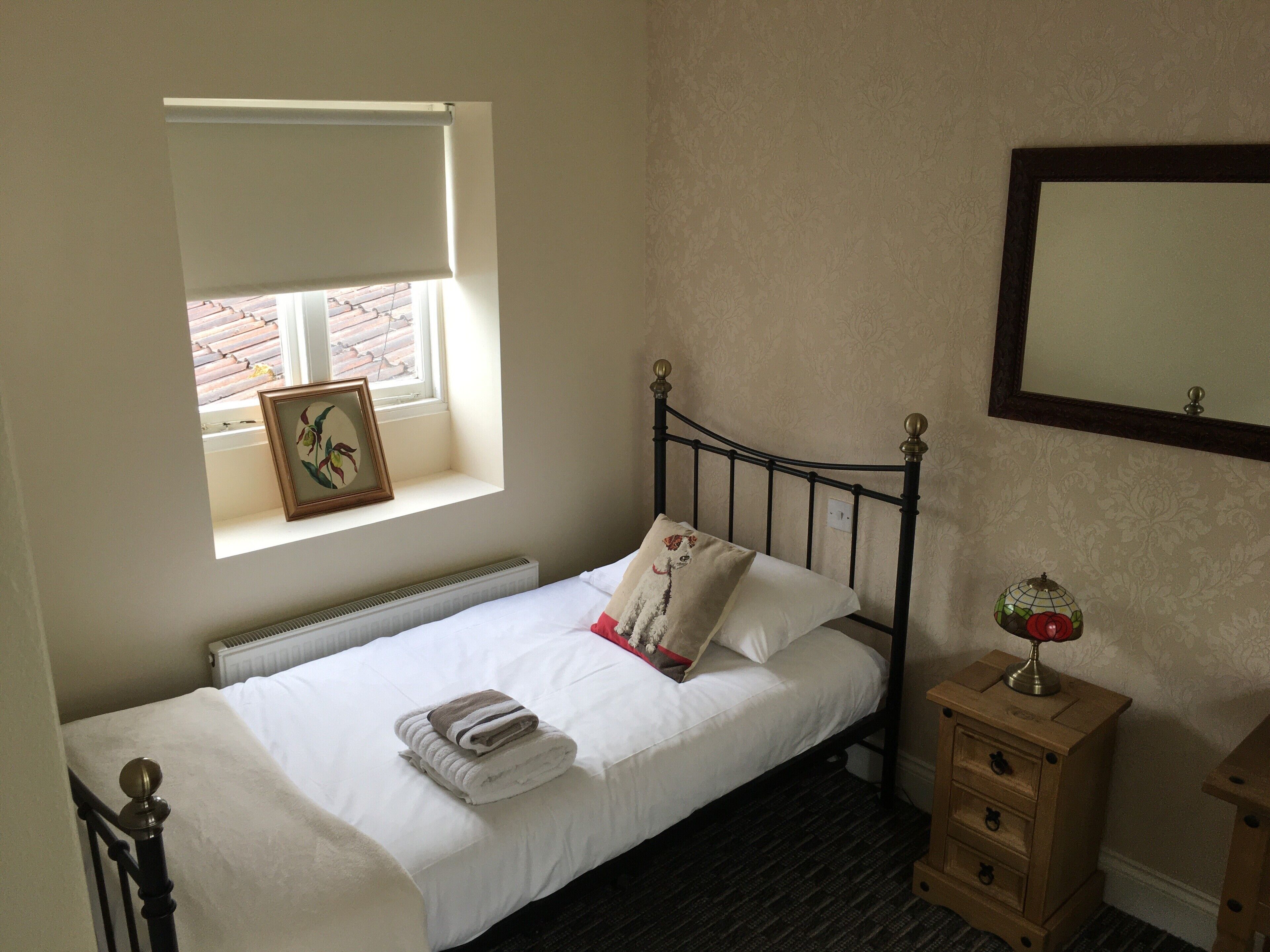 A small, neatly made bed with a white duvet and pillows, a framed picture on the window sill, and a small wooden nightstand with a lamp.