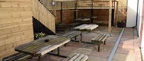 Un patio con bancos y mesas de madera, una estructura de pérgola y un camino de piedra.