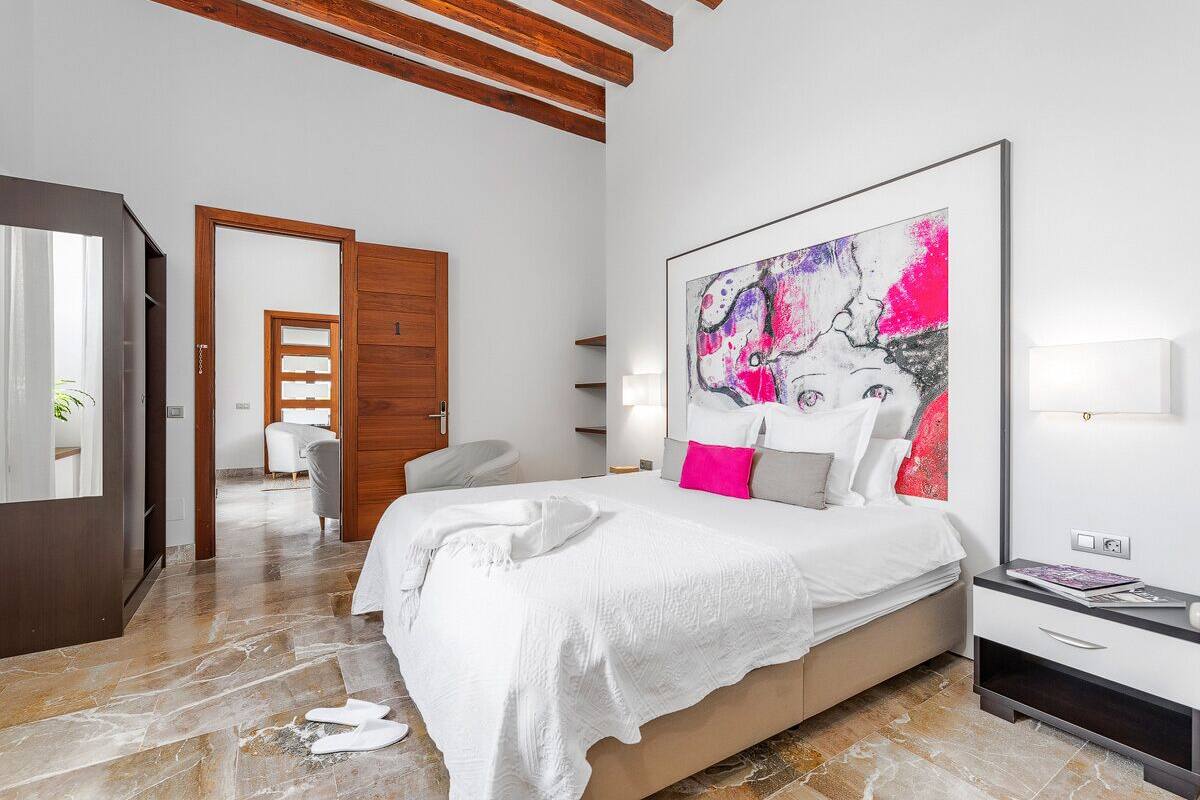 A modern bedroom with a large bed, a wooden ceiling, a large abstract painting, and a bathroom visible in the background.
