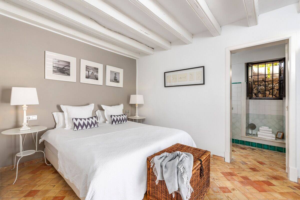 A bedroom with a large bed, two bedside tables with lamps, a wicker bench, and a bathroom visible through an open door.