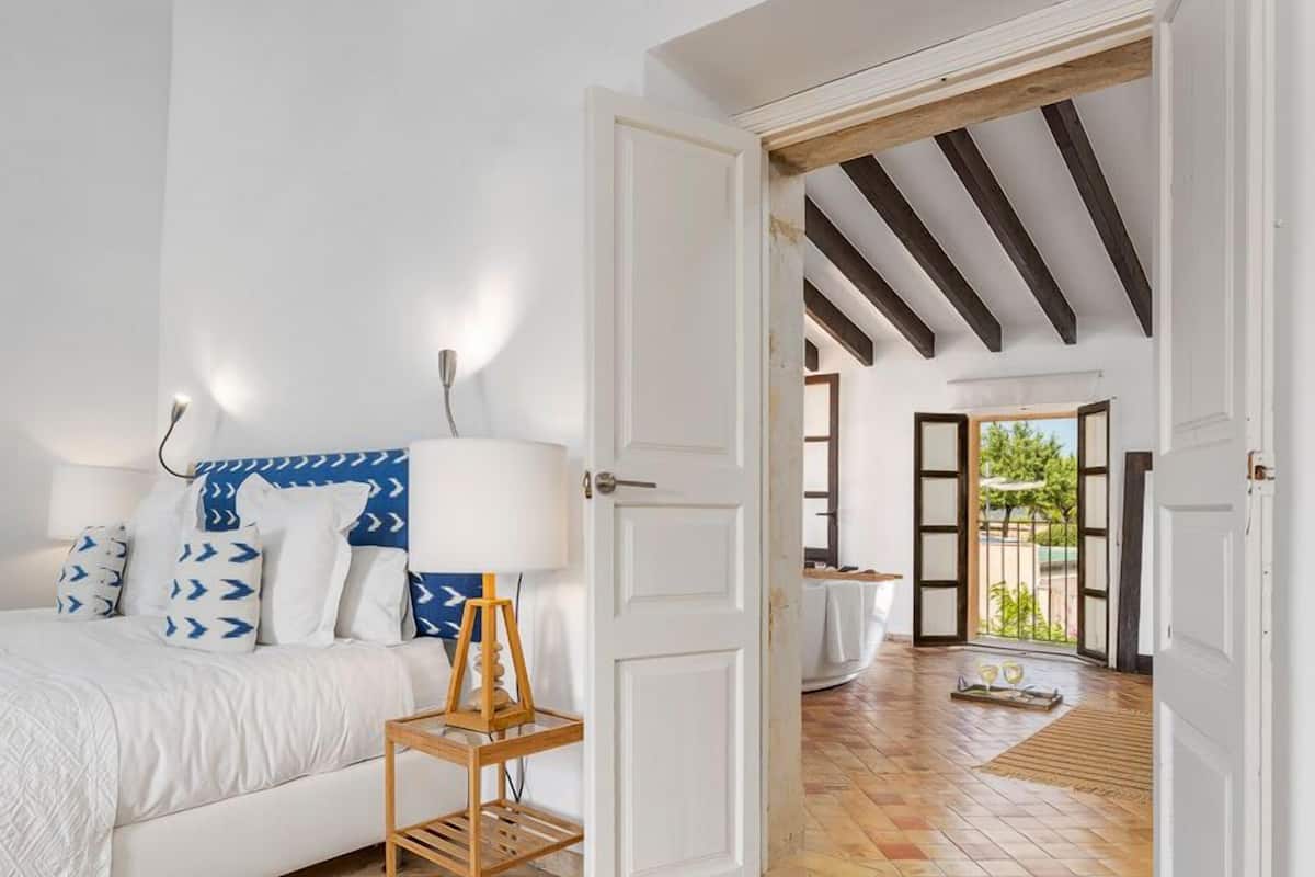 A bedroom with a bed, a bedside table, and a lamp. There is a door leading to another room.