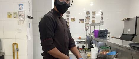 Un chef en una cocina con paredes de azulejos blancos, mostradores de acero inoxidable y diversos utensilios de cocina.
