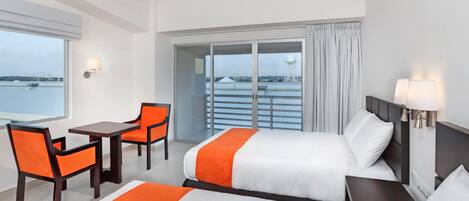 Quarto de hotel com cama, duas cadeiras, uma mesa, luminária e vista para a água pela janela.