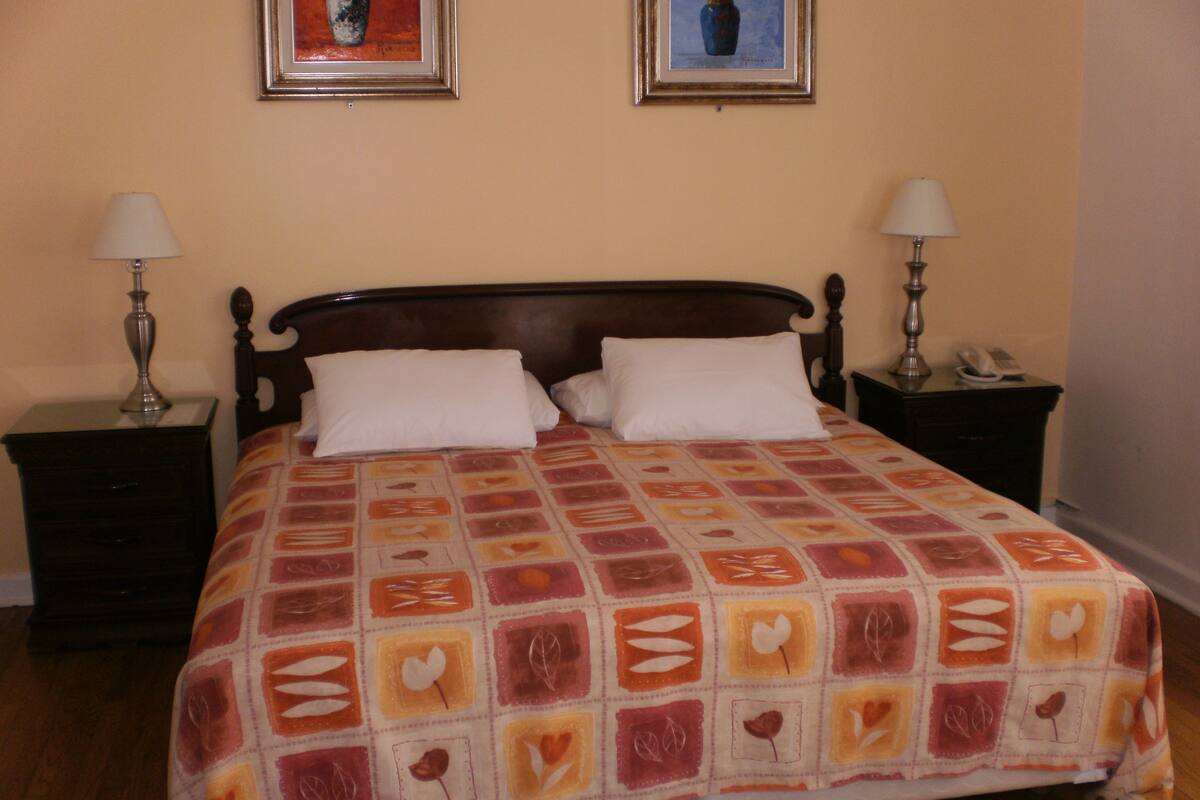 Una cama bien hecha con una colcha estampada, dos mesitas de noche con lámparas, y dos cuadros en la pared.