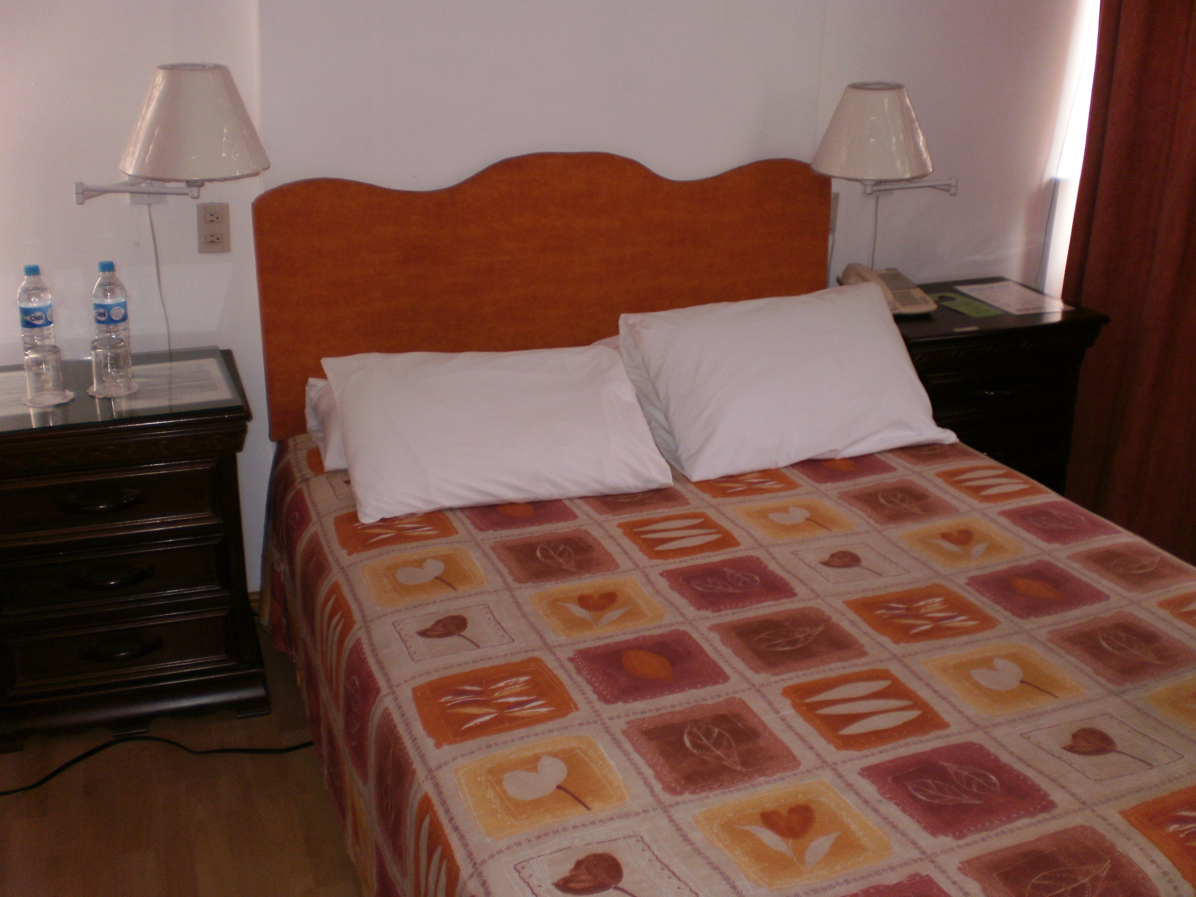 Una cama bien tendida con una colcha estampada, dos lámparas de noche, una mesita con una botella de agua y un aparador de madera.