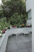 Un espace patio agrémenté d’un banc en métal et d’une petite table, entouré d’une variété de plantes et de fleurs.