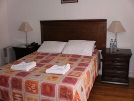 Un lit bien fait, recouvert d’une couvre-lit à motifs, deux lampes de chevet, une table de chevet avec un téléphone et une commode en bois avec des bouteilles d’eau.