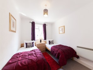 A small, clean room with two beds, a window with curtains, a bedside table, and a radiator.