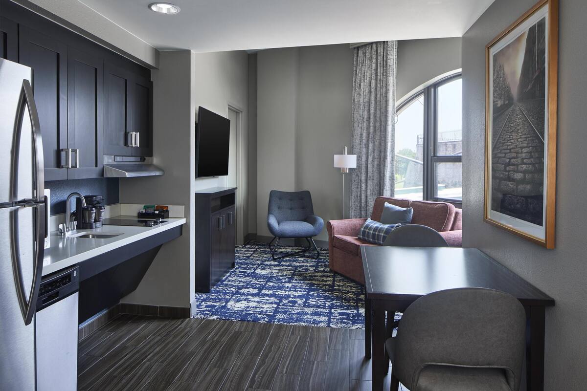 Een compacte hotelkamer met kitchenette, een eethoek, een zithoek met een bank en fauteuil, een groot raam en een schilderij aan de muur.