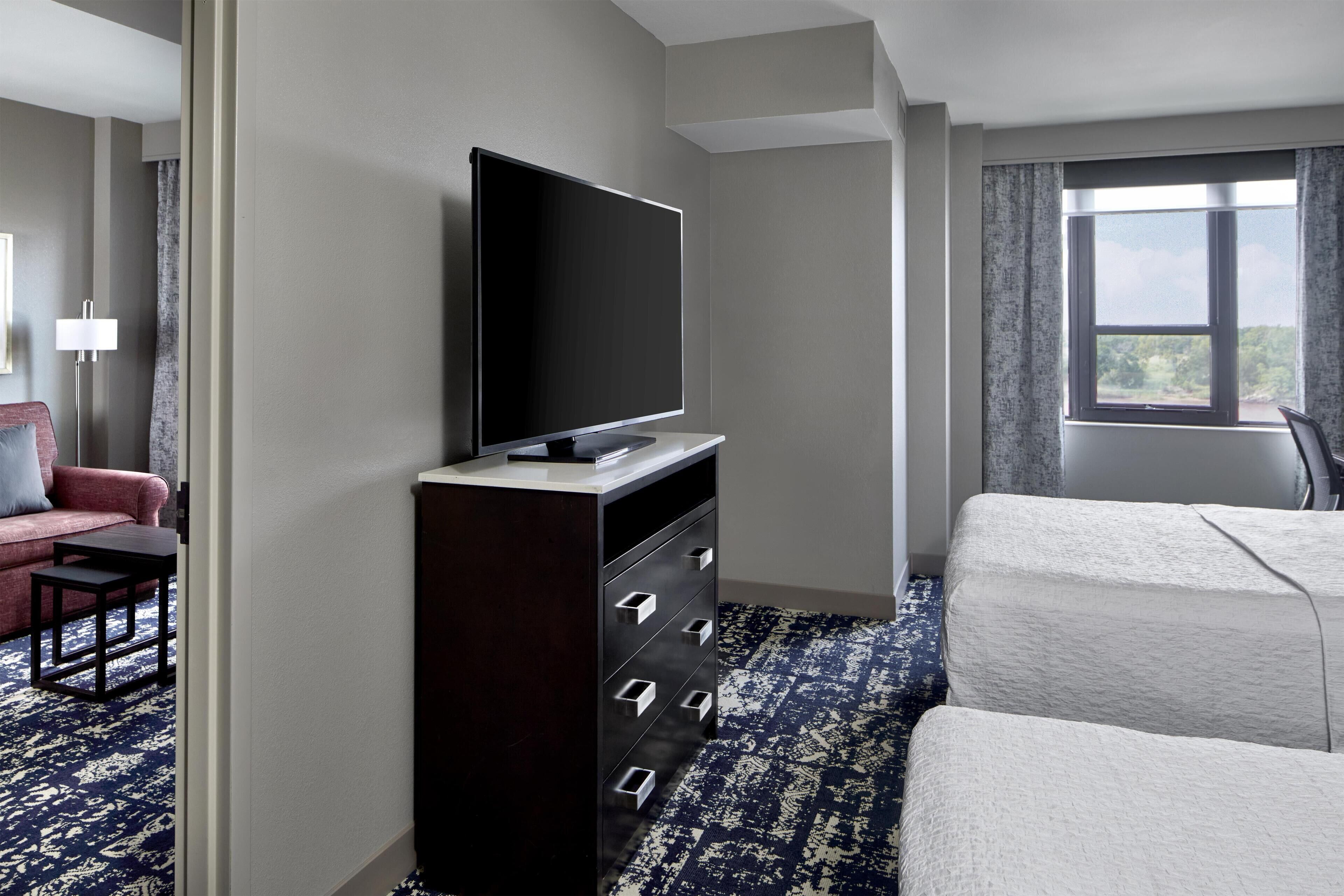 Una habitación de hotel con un televisor de pantalla plana sobre una cómoda, una cama, un ventanal con vistas y un sofá rojo con una mesita.