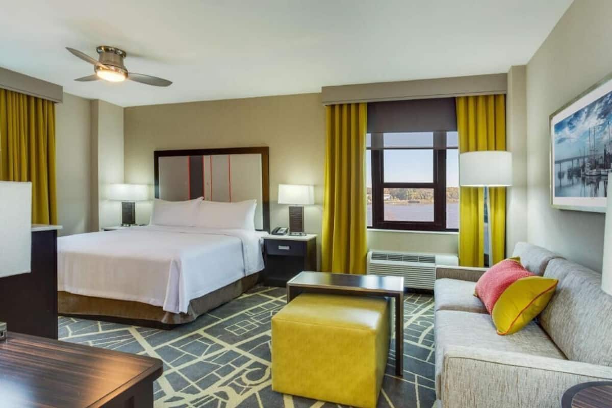 Hotelkamer met een bed, een bank, een kleine tafel, een nachtkastje, een lamp, een plafondventilator en een raam met gordijnen.