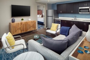A modern hotel room with a sofa, armchairs, a coffee table, a flat-screen TV, a minibar, a bed, and a kitchenette.