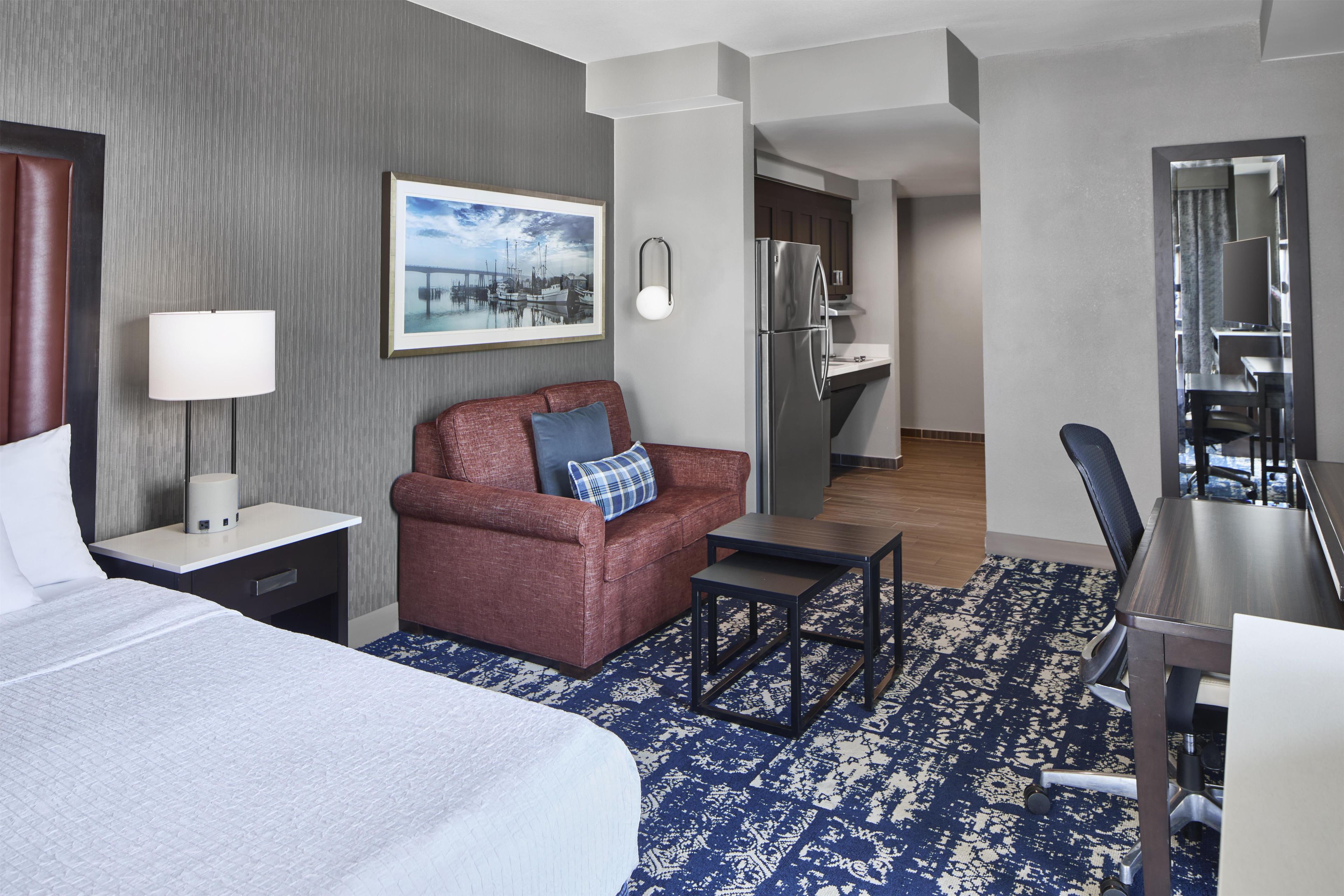 Ein Hotelzimmer mit einem Bett, einem Sofa, einem Schreibtisch, einem Stuhl, einer Lampe, einem Bild an der Wand und einer Küche im Hintergrund.