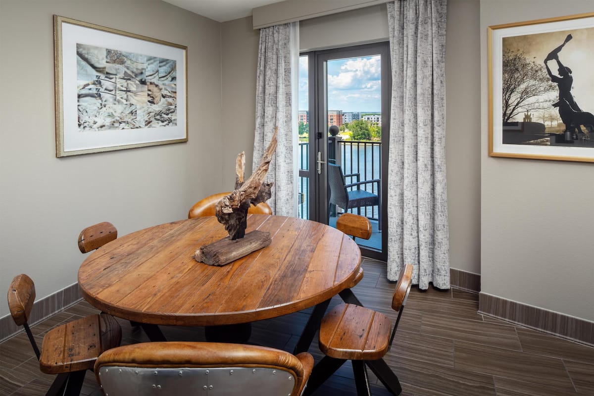 Een ronde houten eettafel met zes stoelen, een groot kunstwerk in een lijst aan de muur, en een balkon met uitzicht op gebouwen en bomen.