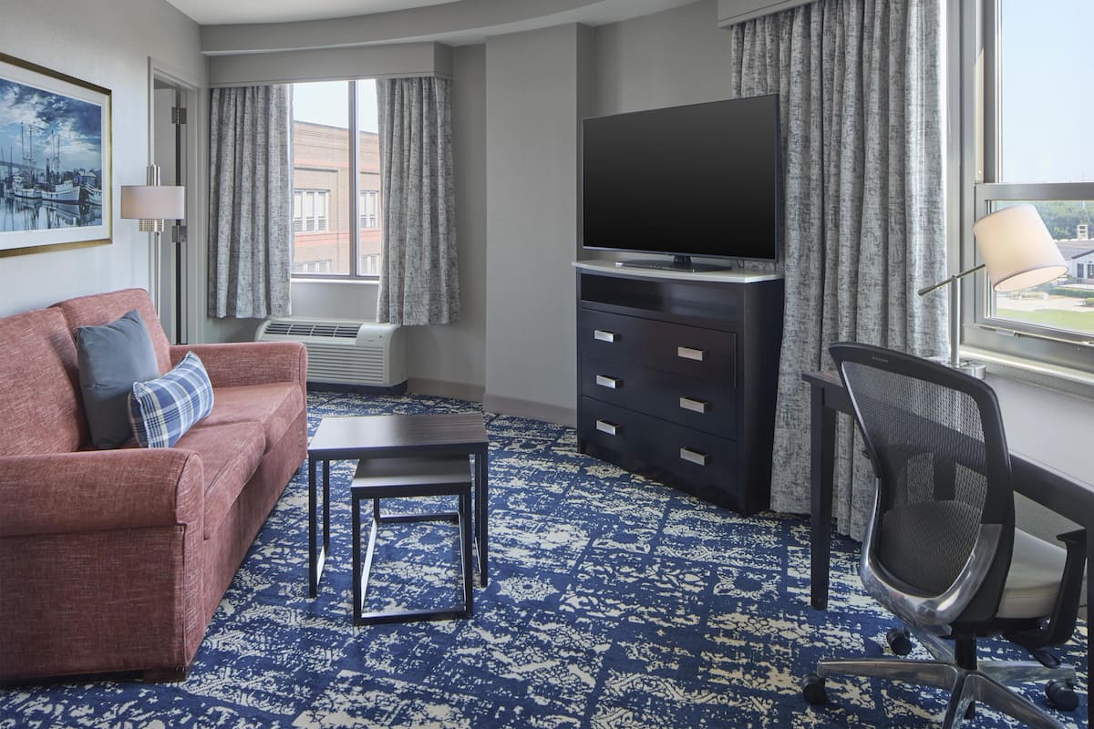 Een hotelkamer met een rode bank, een klein tafeltje, een televisie op een dressoir, een stoel en een groot raam met gordijnen.