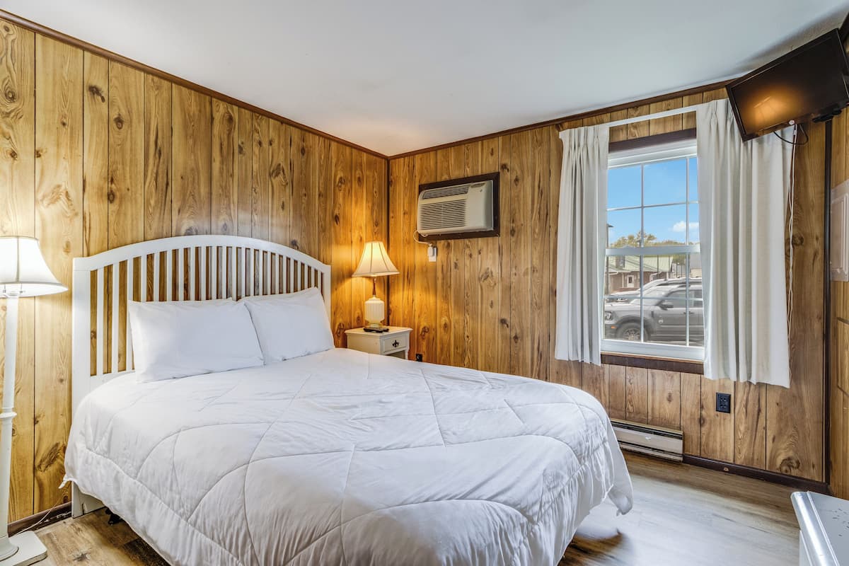 A bedroom with a wooden paneled wall, a bed with a white headboard, a nightstand with a lamp, and a window with curtains.