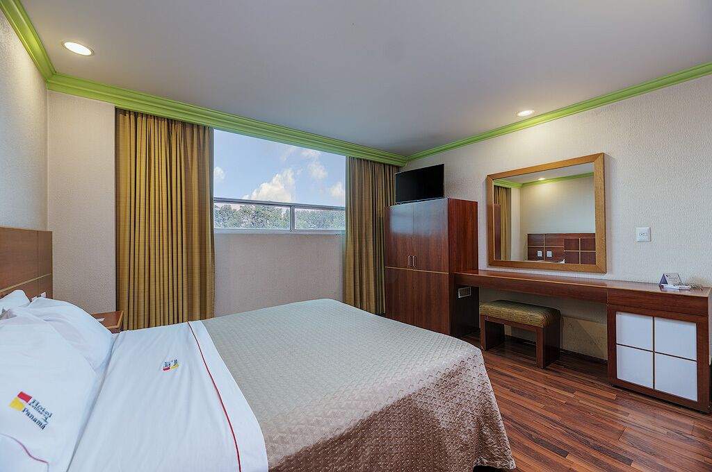 A hotel room with a bed, a desk, a mirror, and a window with curtains.