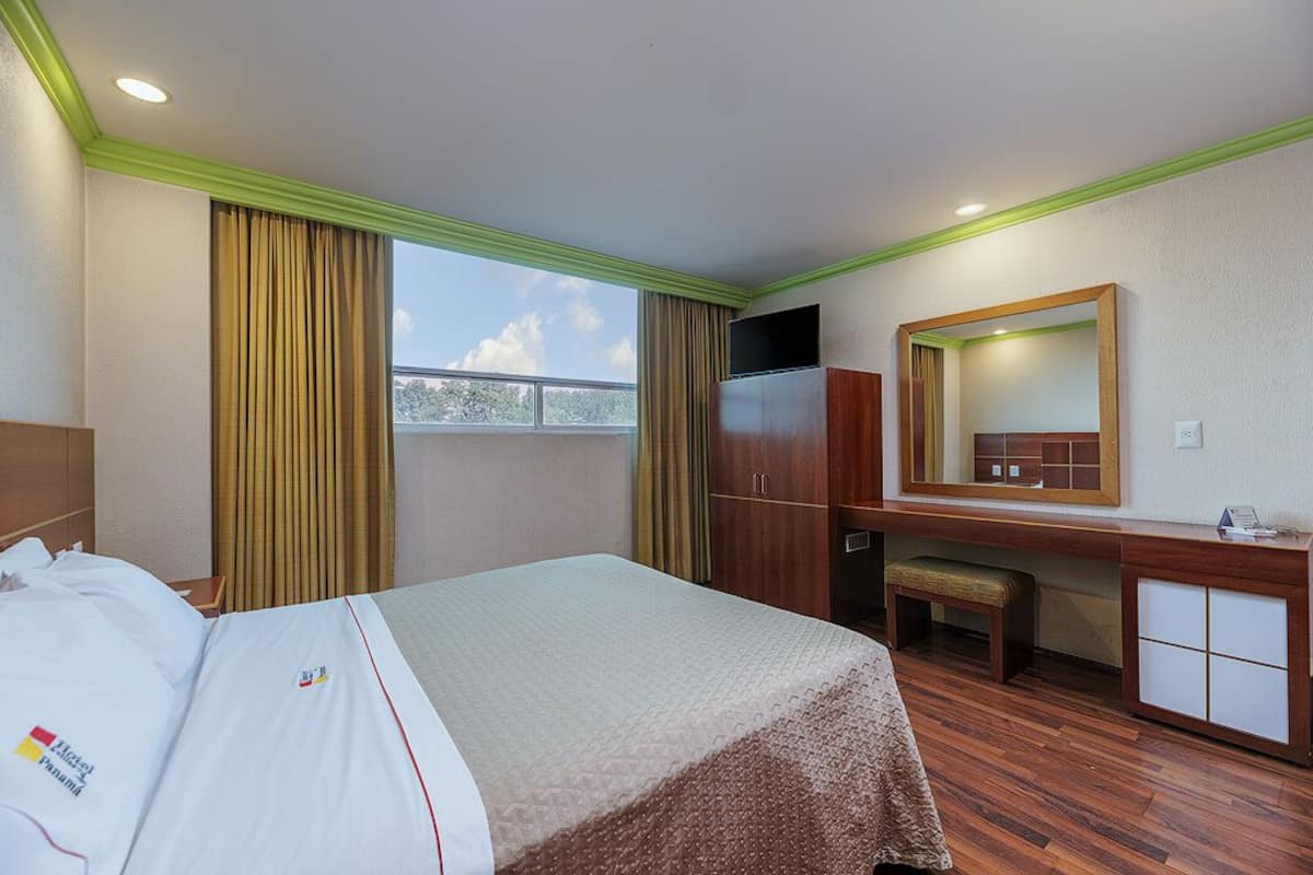 A hotel room with a bed, a desk, a mirror, and a window with curtains.
