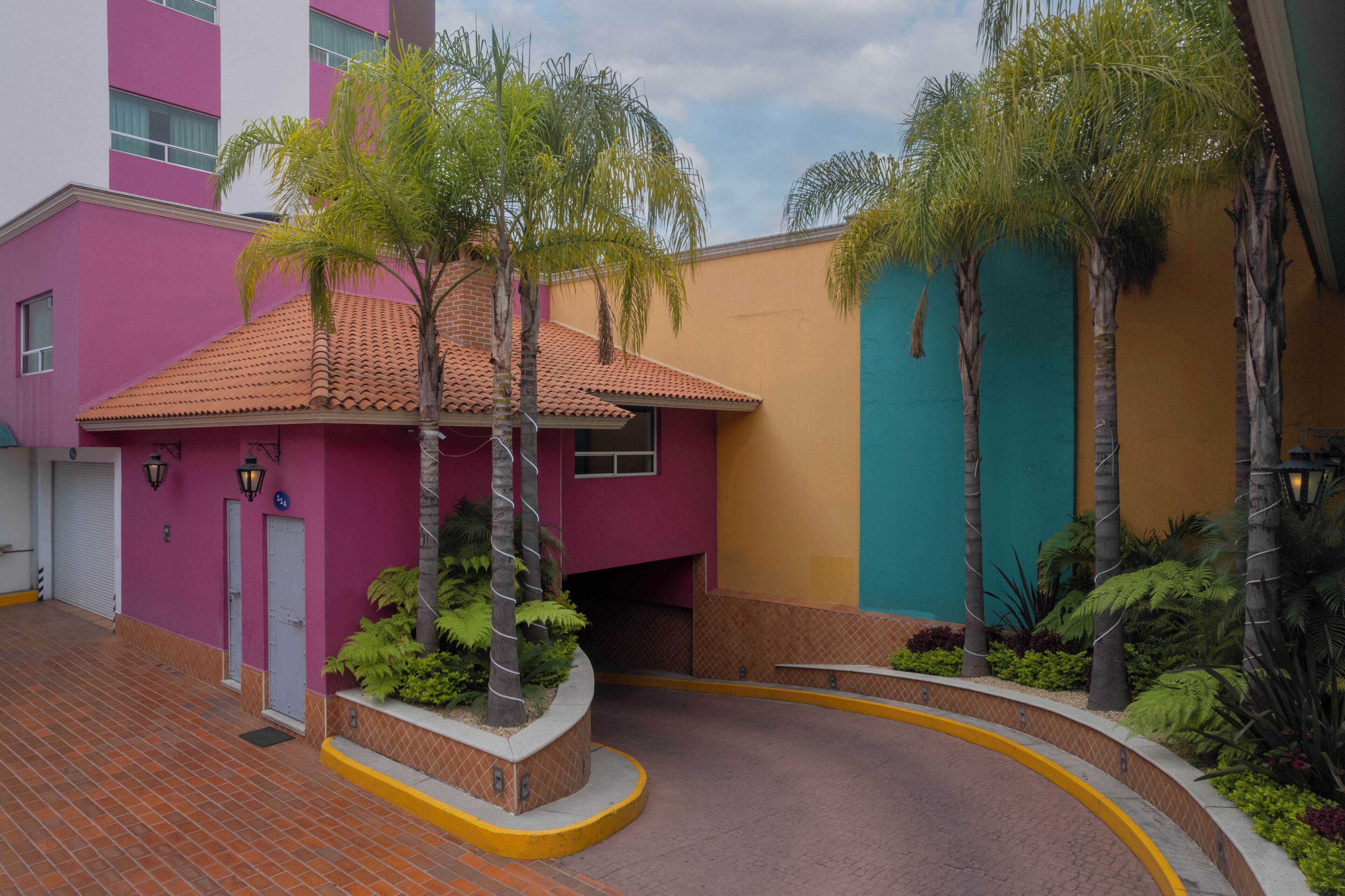 A building with a pink facade, a tiled roof, and a curved pathway with palm trees.