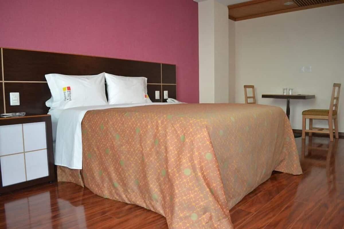 A hotel room with a bed, a bedside table, a chair, and a small table with two glasses.