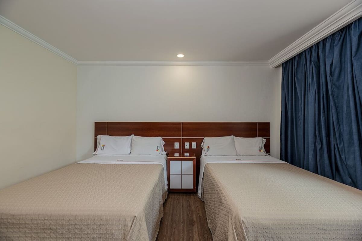 A hotel room with two single beds, a wooden headboard, a nightstand with a lamp, and a large window with blue curtains.