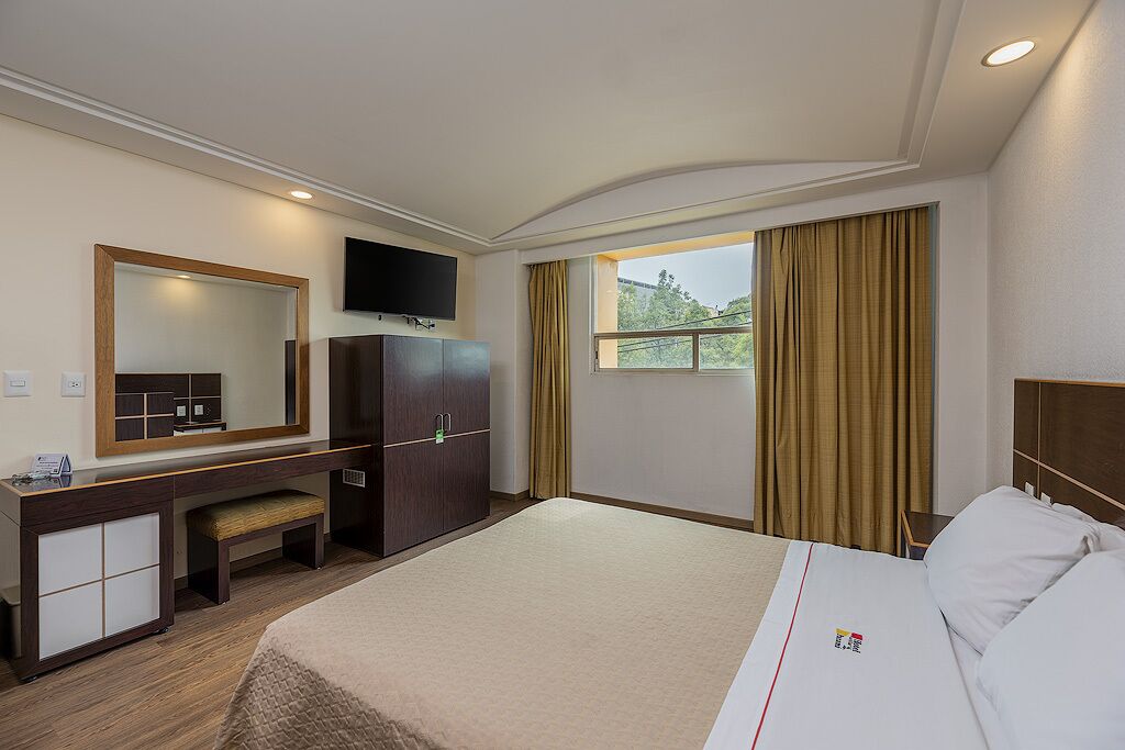 A hotel room with a bed, a desk, a mirror, a television, and a window with curtains.