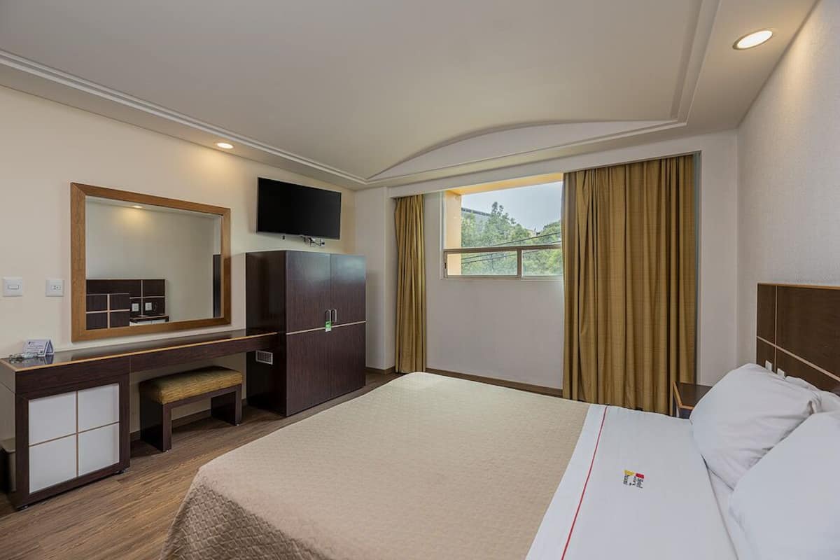 A hotel room with a bed, a desk, a mirror, a television, and a window with curtains.