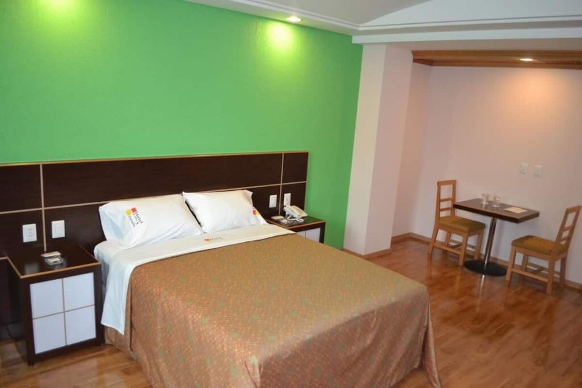 A hotel room with a bed, a bedside table with a phone, a small table with chairs, and a green wall.