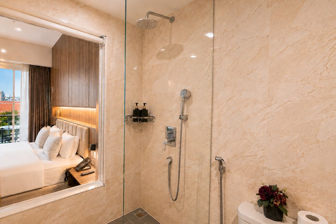 A modern hotel bathroom with a glass-enclosed shower, a toilet with a potted plant, and a bed with a view of the city.