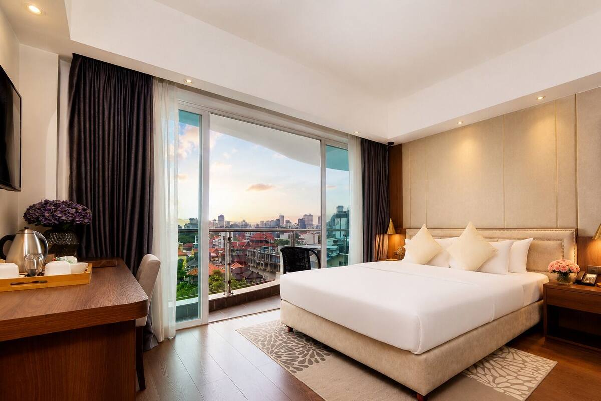 A modern hotel room with a large bed, a desk, a chair, and a view of the cityscape.
