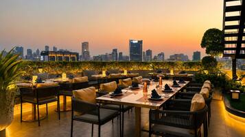 Un espacio de comedor al aire libre con mesas y sillas, un horizonte urbano de fondo y un atardecer.