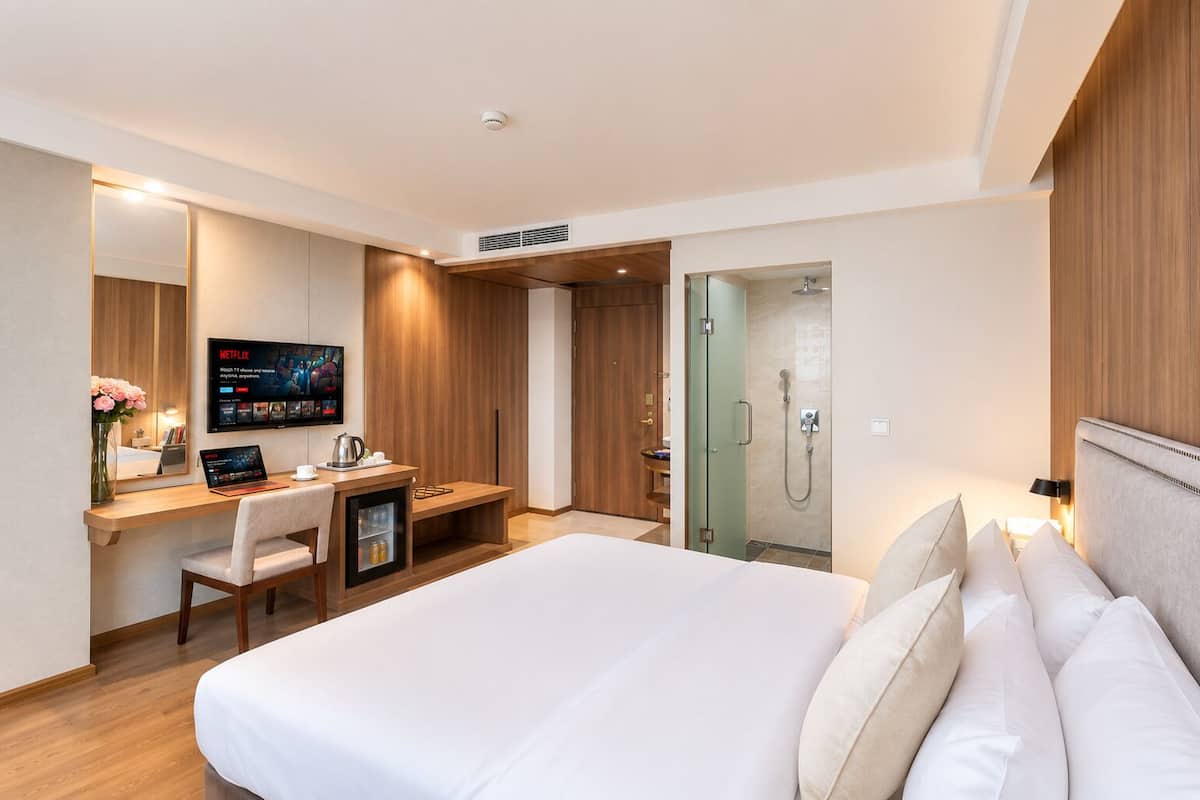 A hotel room with a bed, a desk with a laptop, a TV, a shower, and a mirror.