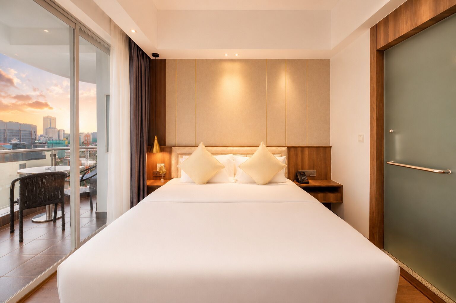 Quarto de hotel com uma cama grande, vista para a cidade, varanda com cadeiras e porta de madeira.