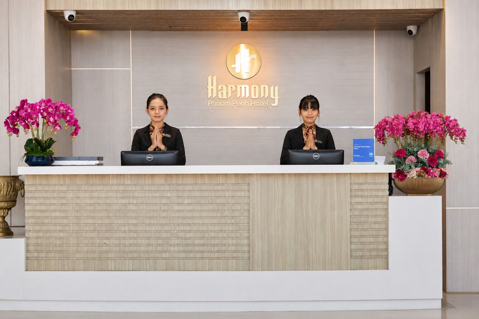 Un comptoir d’accueil à l’hôtel Harmony Phnom Penh, avec deux réceptionnistes, un panneau indicateur et des plantes en pot.