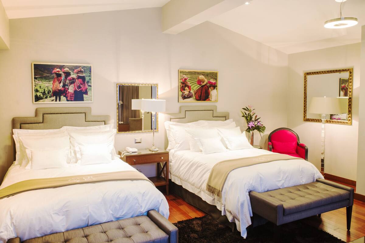 A hotel room with two beds, a red chair, a bedside table with a lamp, and two paintings on the wall.